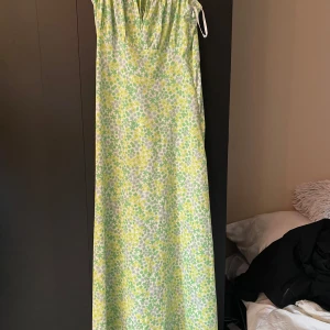 Blommig långklänning med smala axelband - Säljer en söt långklänning med grönt och gult blommönster. Klänningen har smala axelband som knyts i ryggen och en lätt v-ringning framtill. Perfekt för varma dagar och somriga tillfällen.