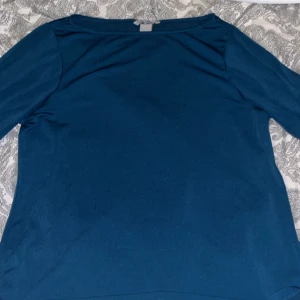 Petrolblå långärmad topp från H&M - Säljer en stilren petrolblå kortärmad topp från H&M. Toppen har en enkel design med diskreta sömmar och lätt puffade ärmar med breda muddar. Materialet känns mjukt och har en lätt glans. Perfekt för dig som gillar minimalistisk stil.