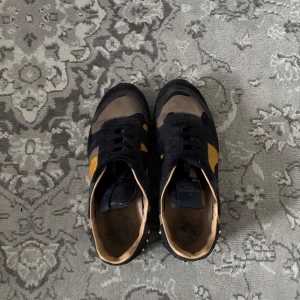 Valentino Rockrunner - Snygga Valentino Rockrunner sneakers med kamouflagemönster i svart, grått och gult. Skorna har svarta snören, nitar på hälen och en robust sula. Märkeslogga på plösen och insidan. Perfekta för dig som vill sticka ut med stil. Liten defekt två nitar saknas.