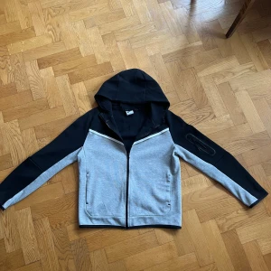 Nike Tech Hoodie - Snygg hoodie från Nike i svart och grått med hel dragkedja framtill. Jackan har huva, två fickor fram och en extra ficka med dragkedja på ärmen. Perfekt för en sportig stil. Kontakta vid minsta fråga 🤗