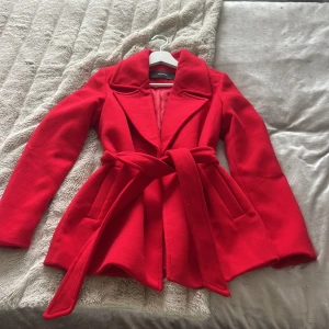 Röd kappa med bälte från Vero Moda - Säljer en snygg röd kappa från Vero Moda med markerad krage och knytskärp i midjan. Jackan har långa ärmar och är helfodrad. Perfekt för dig som vill sticka ut med färg i höst och vinter.
