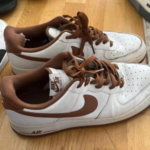 Nike Air Force 1 sneakers i vitt och brunt - Nike Air Force 1 sneakers i vitt med bruna detaljer, swoosh och snörning. Klassisk modell med perforerad tå och låg profil. Bekväm innersula och platt sula. Perfekta för dig som gillar retrostil och streetwear. De är på acceptabelt skick men, är sönder bara vid hälarna. Har köpt de i JD sport för 1300kr och säljer de för 400kr ( vid snabb köp kan priset diskuteras)