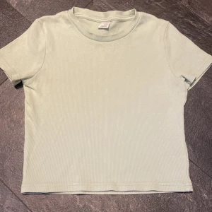 Ljusgrön ribbad t-shirt från Lindex - En ljusgrön ribbad t-shirt från Lindex med rund halsringning och korta ärmar. T-shirten har en enkel och stilren design, perfekt för en avslappnad look. Materialet är mjukt och stretchigt för extra komfort.