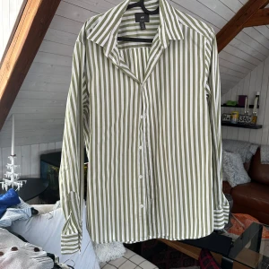 Grön- och vitrandig skjorta från H&M - Säljer en grön- och vitrandig skjorta från H&M i regular fit. Skjortan har långa ärmar, klassisk krage och knäppning framtill. Perfekt för dig som gillar en stilren och avslappnad look.storlek s
