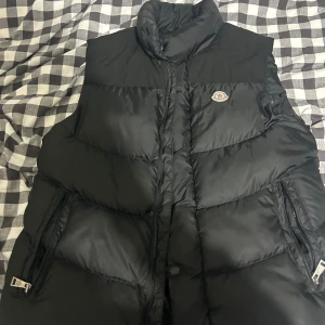 Svart dunväst från Moncler - Säljer en svart dunväst från Moncler med hög krage och dragkedja framtill. Västen har två fickor med dragkedja och klassisk Moncler-logga på bröstet. Perfekt för lager-på-lager och stilren look.