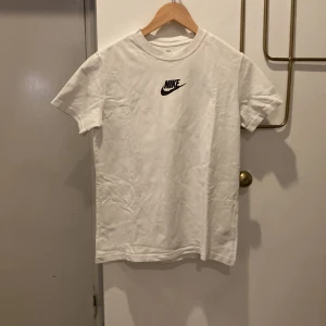 Vit t-shirt från Nike - Säljer en klassisk vit t-shirt från Nike med svart logga på bröstet. T-shirten har rund halsringning och korta ärmar. Perfekt basic-plagg med normal passform och tillverkad i mjuk bomull.