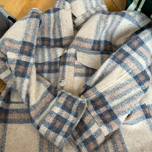 Beige och blå rutig kappa från H&M - Säljer en snygg kappa från H&M i beige med blått och brunt rutmönster. Jackan har långa ärmar, stora knappar framtill och klassisk krage. Perfekt för dig som gillar en stilren och trendig look.