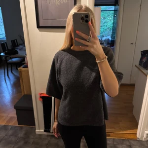 Mörkgrå stickad tröja med kort ärm - Mysig mörkgrå stickad tröja med rund halsringning och korta ärmar. Tröjan har en rak och lite loose passform, perfekt för lager-på-lager-stil. Enkel och stilren design som passar till mycket.