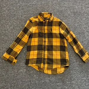 Gul och svart rutig skjorta från Next - Säljer en snygg gul och svart rutig skjorta från Next i 100% bomull. Skjortan har långa ärmar, klassisk krage och knäppning framtill. Perfekt för dig som gillar färg och mönster!