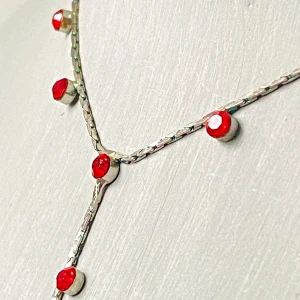 Silverfärgat halsband med röda stenar - Ett elegant halsband i silverfärgad metall med små röda stenar jämnt placerade längs kedjan. Halsbandet har en Y-formad design som ger en stilren och modern look. Perfekt för att addera färg till din outfit.