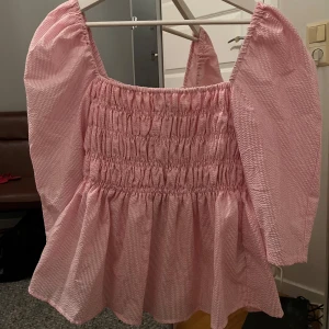 Rosa blus med puffärmar från VILA - Supersöt rosa blus från VILA med smockad överdel och puffiga långärmar. Blusen har ett diskret vitt randigt mönster och volang nedtill. Perfekt för dig som gillar romantisk stil och vill ha något extra till garderoben!