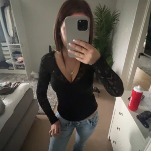 Svart spetsblus med v-ringning - Säljer en svart blus med v-ringning och långa ärmar i spets. Sitter tight och har ett elegant, genombrutet mönster på ärmarna. Perfekt att styla med jeans eller kjol.