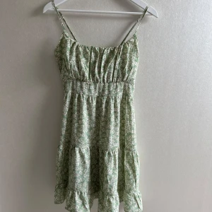 Grön blommig sommarklänning med volang - Söt ljusgrön klänning med vita blommor och smala justerbara axelband. Klänningen har volang nertill och smock i ryggen för en bekväm passform. Perfekt till sommaren!💗💗