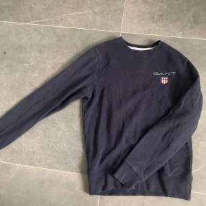 Mörkblå sweatshirt från GANT - Säljer en mörkblå sweatshirt från GANT med rund halsringning och broderad logga på bröstet. Tröjan har långa ärmar och ribbade muddar vid ärmslut och nederkant. Perfekt för en avslappnad stil.