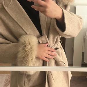 Beige kappa med fuskpälsdetaljer och bälte - Säljer en beige kappa med stora fuskpälsdetaljer vid krage och ärmslut. Kappan har ett knytbälte i midjan och en klassisk, elegant design. Perfekt för dig som vill ha en stilren och mysig jacka till kyligare dagar.