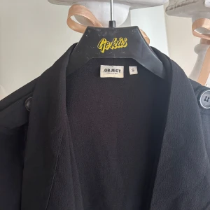 Svart trenchcoat från Object - Säljer en svart trenchcoat från Object i storlek S. Jackan har långa ärmar, bred krage och snygga detaljer vid axlarna. Perfekt för dig som vill ha en stilren och tidlös jacka till garderoben.