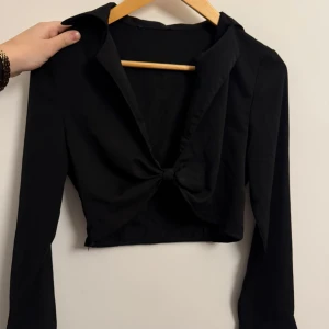 Svart croppad blus med knyt - Säljer en svart croppad blus med lång ärm och snygg knytning framtill. Perfekt för dig som vill ha en trendig och stilren topp med krage och djup v-ringning.