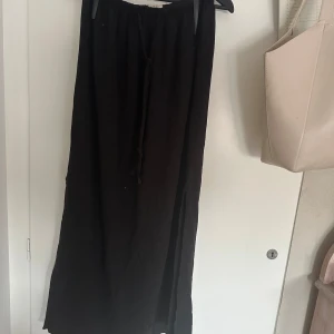 Svart långkjol från Gina Tricot - Svart långkjol från Gina Tricot med resår i midjan och knytband. Kjolen har en slits på sidan som ger en snygg detalj. Fint skick som inte är använd så många gånger, ca 3 gångers användning. Förlåt för lite dåliga bilder! Skriv vid intresse eller fundering!❤️🙏