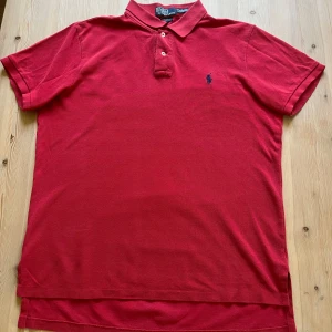 Röd pikétröja från Polo Ralph Lauren - Klassisk röd pikétröja från Polo Ralph Lauren med blå broderad logga på bröstet. Tröjan har krage, två knappar och är tillverkad i bomull. Perfekt för en stilren och sportig look. Size L