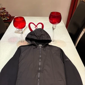 Svart jacka med huva från Barbour - Säljer en svart jacka från Barbour med huva och dragkedja. Jackan har en stilren design och är tillverkad i ett syntetmaterial. Perfekt för dig som gillar enkel och funktionell stil.