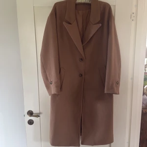 Beige kappa från Divided - Säljer en klassisk beige kappa från Divided med lång passform och stora slag. Kappan har två knappar framtill och fickor på sidorna. Perfekt för höst och vår, enkel att matcha till olika outfits. Stor i storlek Ca M! 