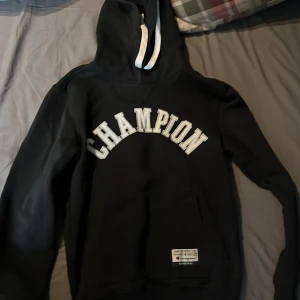 Svart hoodie från Champion - Svart hoodie från Champion med vit text över bröstet och vit dragsko i huvan. Klassisk känguruficka framtill och ribbade muddar vid ärmslut och nederkant. Perfekt för en avslappnad stil.