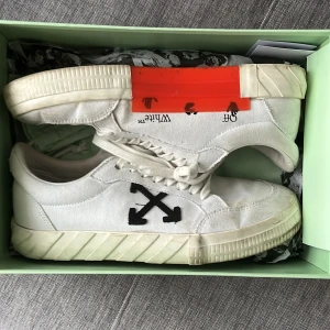 Vita sneakers från Off-White - Säljer ett par vita sneakers från Off-White med svart pil-logga på sidan och en tydlig orange detalj på sulan. Skorna har snörning och är tillverkade i canvas med gummisula. Perfekta för dig som gillar streetwear och vill sticka ut med en unik design. Helt ok skick 5/10. Försöker bara bli av med skorna! 