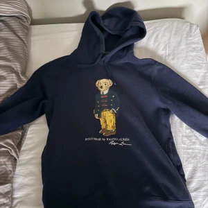 Marinblå hoodie med Polo Bear från Ralph Lauren - Säljer en marinblå hoodie från Ralph Lauren med Polo Bear-tryck på bröstet. Tröjan har huva med dragsko och klassisk passform. Perfekt för dig som gillar streetwear och vill ha något stilrent men ändå lekfullt. Nypris runt 2.5 och denna är i absolut nyskick. Använd 1 gång kanske 