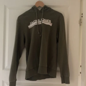 Olivgrön hoodie från Jack & Jones - Säljer en olivgrön hoodie från Jack & Jones med vit logga på bröstet. Tröjan har huva med dragsko och långa ärmar. Perfekt för en avslappnad stil.
