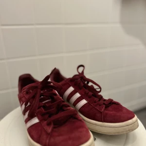 Vinröda Adidas Gazelle sneakers - Säljer ett par vinröda Adidas Gazelle sneakers med klassiska vita ränder på sidorna och vit sula. Skorna har snörning och är tillverkade i mocka. Kommer givetvis tvättas extra innan köp