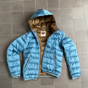 Blå pufferjacka från Colmar - Snygg blå pufferjacka från Colmar med brunt foder och huva. Jackan har dragkedja framtill, två fickor med dragkedja och en patch med Colmar-logga på ärmen. Två sista bilderna visar defekterna som finns på jackan. Storlek XS-S.  Skriv till mig om du har funderingar. 