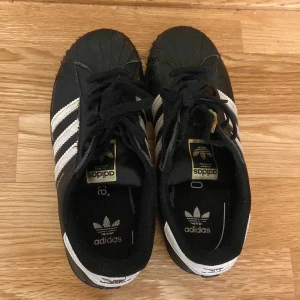 Adidas Superstar svartvita sneakers - Klassiska Adidas Superstar sneakers i svart med vita ränder och vit häl. Skorna har snörning och den ikoniska gummitån framtill. Adidas-logga på plös och häl. Perfekta för en sportig och stilren look.