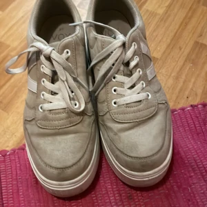 Beige sneakers med platåsula från VOX - Säljer ett par beige sneakers från VOX med vit platåsula och snörning. Skorna har en stilren design med diskreta detaljer och en vit rand på sidan. Perfekta för dig som vill ha lite extra höjd och en modern look.