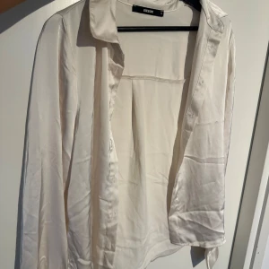 Vit satinblus från Bik Bok - Säljer en stilren creme vit blus från Bik Bok i glansigt satinmaterial. Skjortan har klassisk krage, långa ärmar med manschett och knappar framtill. Perfekt att styla till olika looks! (Aldrig använd, kommer tyvärr inte till användning) 