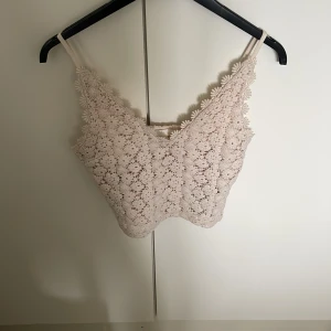 Beige spetslinne med blommönster - Säljer ett superfint beige linne med smala axelband och vackert blommigt spetsmönster. Toppen är croppad och har en romantisk känsla, perfekt för sommardagar eller som ett lager under en skjorta.