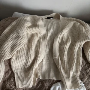 Beige stickad kofta - Säljer en beige stickad kofta med öppen front och ribbade muddar. Koftan har lång ärm och är perfekt för lager-på-lager-stil. Enkel och stilren design som passar till mycket.