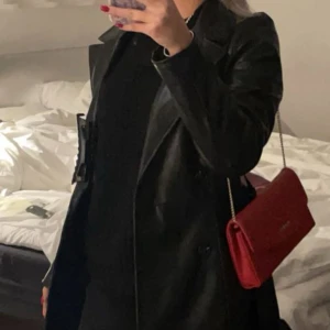 Svart skinn trenchcoat - Snygg svart trenchcoat i skinn med klassisk krage och knäppning framtill. Jackan har en stilren och tidlös design som passar perfekt till många olika outfits. Kombinera med en färgglad väska för extra statement.