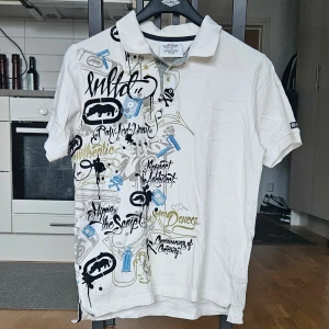 Vit piké t-shirt med tryck från Ecko Unltd. - Vit piké t-shirt från Ecko Unltd. med coolt grafiskt tryck i svart, blått och gult på både fram- och baksidan. Klassisk krage och korta ärmar. 100% bomull, perfekt för en streetwear-look.