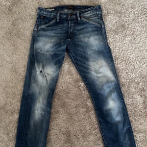 Blå slitna jeansbyxor från Jack & jones - Säljer ett par blå jeansbyxor från Replay med slitningar och ljusa tvättade partier. Klassisk femficksmodell med knappgylf och läderpatch bak i midjan. Perfekta för en avslappnad stil. Slim fit passform storlek W29 L32, litet hål vid knät:)
