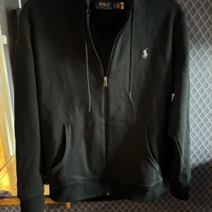 Polo Ralph Lauren zip  - Säljer en svart hoodie från Polo Ralph Lauren med dragkedja och huva. Tröjan har två fickor framtill och den klassiska broderade loggan i vitt på bröstet. Perfekt för en avslappnad stil. Använts någongång men inget som syns på den:) storlek M