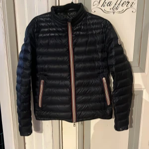 Mörkblå pufferjacka från Moncler - Säljer en mörkblå pufferjacka från Moncler med klassisk quiltad design. Jackan har lång ärm, ståkrage och dragkedja framtill samt vid fickorna. Snygga röd-vita detaljer på dragkedjorna och Moncler-logga på ärmen.