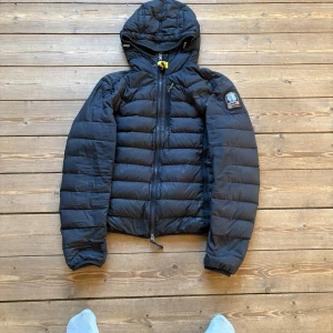 Svart dunjacka med huva från Parajumpers - Säljer en svart dunjacka från Parajumpers med luva och dragkedja. Jackan är superlätt och har en lapp på ärmen. Klassisk pufferstil med quiltad design och snygga detaljer som ett gult spänne i nacken. Observera att jackan är använd ganska mycket, och att den därför saknar dragkedja och har tecken på slitage som hål och fläckar som du inte kommer att märka så mycket av.