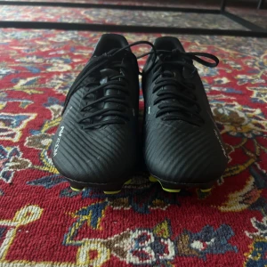  Nike Mercurial fotbollsskor - Säljer ett par svarta Nike Mercurial fotbollsskor med snörning och gula dobbar. Skorna har en sportig design med rund tå och platt sula, perfekta för spel på gräsplan (går att spela på konstgräs med men slits lite snabbare). Snygg och stilren look med diskreta gråa detaljer och Mercurial-logga på hälen. Knappt använt som nya.