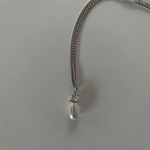 Silverfärgat halsband med pärlhänge från Marianilsdotter - Säljer ett stilrent silverfärgat halsband från Marianolsdotter  med ett droppformat pärlhänge. Kedjan är klassisk och smal, och hänget har en liten dekorativ detalj upptill. Perfekt för dig som gillar enkel och elegant design.