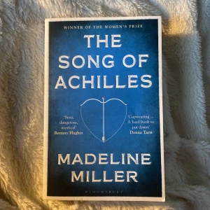 The Song of Achilles - En hyllad roman av Madeline Miller som återberättar den grekiska myten om Akilles och Patroklos. Boken har ett blått omslag med ett hjärta och pil, och är skriven på engelska. Perfekt för dig som gillar historiska romaner och mytologi.