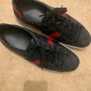 Svarta sneakers från Gucci med röd och blå rand - Säljer ett par svarta Gucci sneakers med klassiskt GG-mönster, röd och blå rand på sidan samt röd detalj vid hälen. Skorna har svarta skosnören och vit sula. Perfekta för dig som gillar lyxiga och stilrena sneakers.
