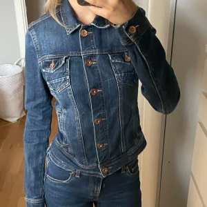 Mörkblå jeansjacka från Bershka - Säljer en klassisk mörkblå jeansjacka från Bershka med bruna knappar och kontrastsömmar. Jackan har två bröstfickor med lock och knapp samt en figurnära passform. Perfekt att slänga över en t-shirt för en cool look. Liten i storleken