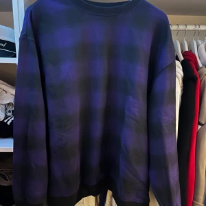 Weekday Lila och svart rutig sweatshirt - Säljer en oversized sweatshirt i lila och svart rutigt mönster från Weekday. Tröjan har rund halsringning och långa ärmar. Perfekt för dig som gillar streetwear och vill ha en bekväm och snygg stil.
