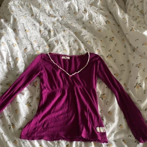Lila långärmad topp från Odd Molly - Säljer en lila långärmad topp från Odd Molly med v-ringning, vit spetskant och en liten rosett framtill. Toppen har broderad text på sidan och är tillverkad i ett ribbat material. Perfekt för dig som gillar unika detaljer!