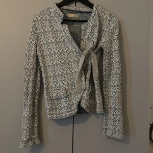 Mönstrad stickad jacka med knyt från Odd Molly - Supersnygg stickad jacka från Odd Molly i vitt och blått med grafiskt mönster. Jackan har lång ärm, två fickor framtill och knytband i midjan. 